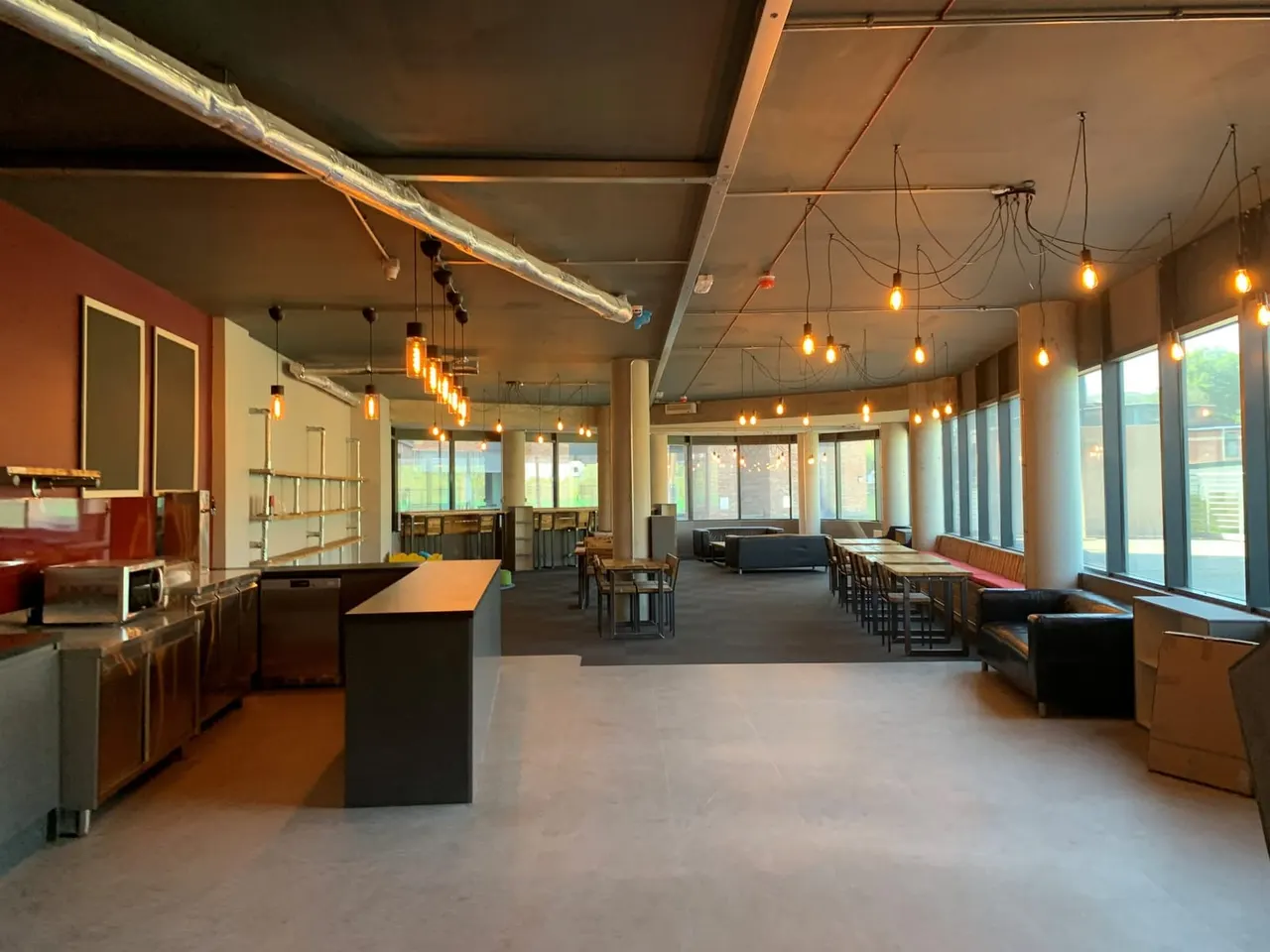 Modern café interior with pendant lighting, kitchen area on left, scattered seating and tables, and large windows along the right side.