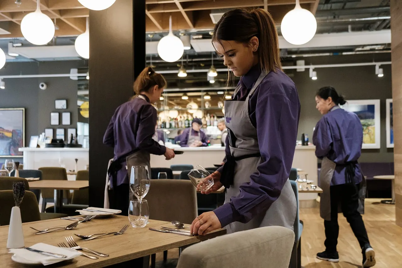 Staff member in purple uniform and apron setting a dining table in a modern restaurant with pendant lights and exposed ceiling.
