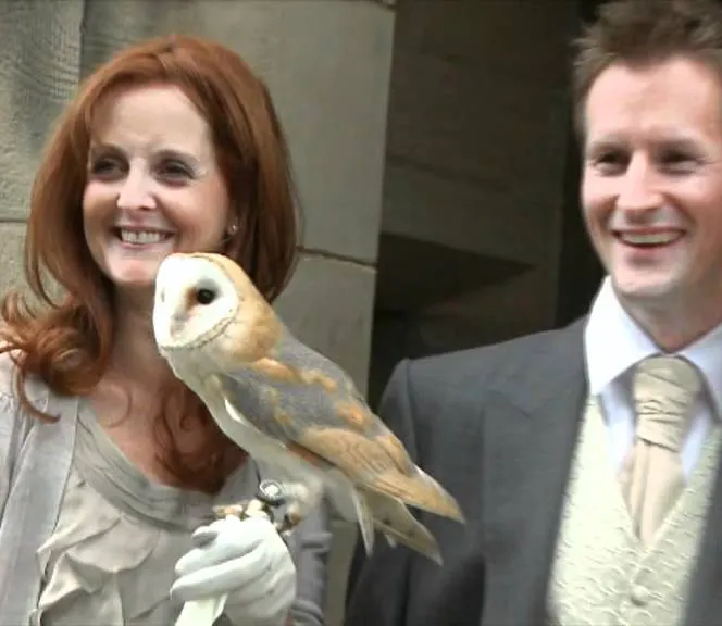 A woman and man in smart casual attire pose whilst holding a barn owl on a gloved hand.