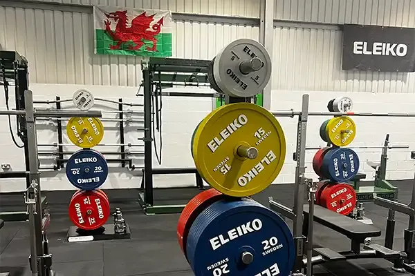 Weightlifting gym interior with coloured barbells and weight plates loaded on racks, Welsh flag visible.