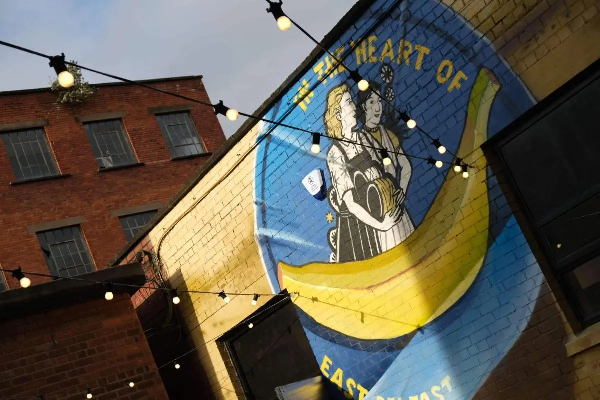 Large mural on brick building showing two figures in a giant yellow banana, with "HEART OF" text visible and string lights overhead.