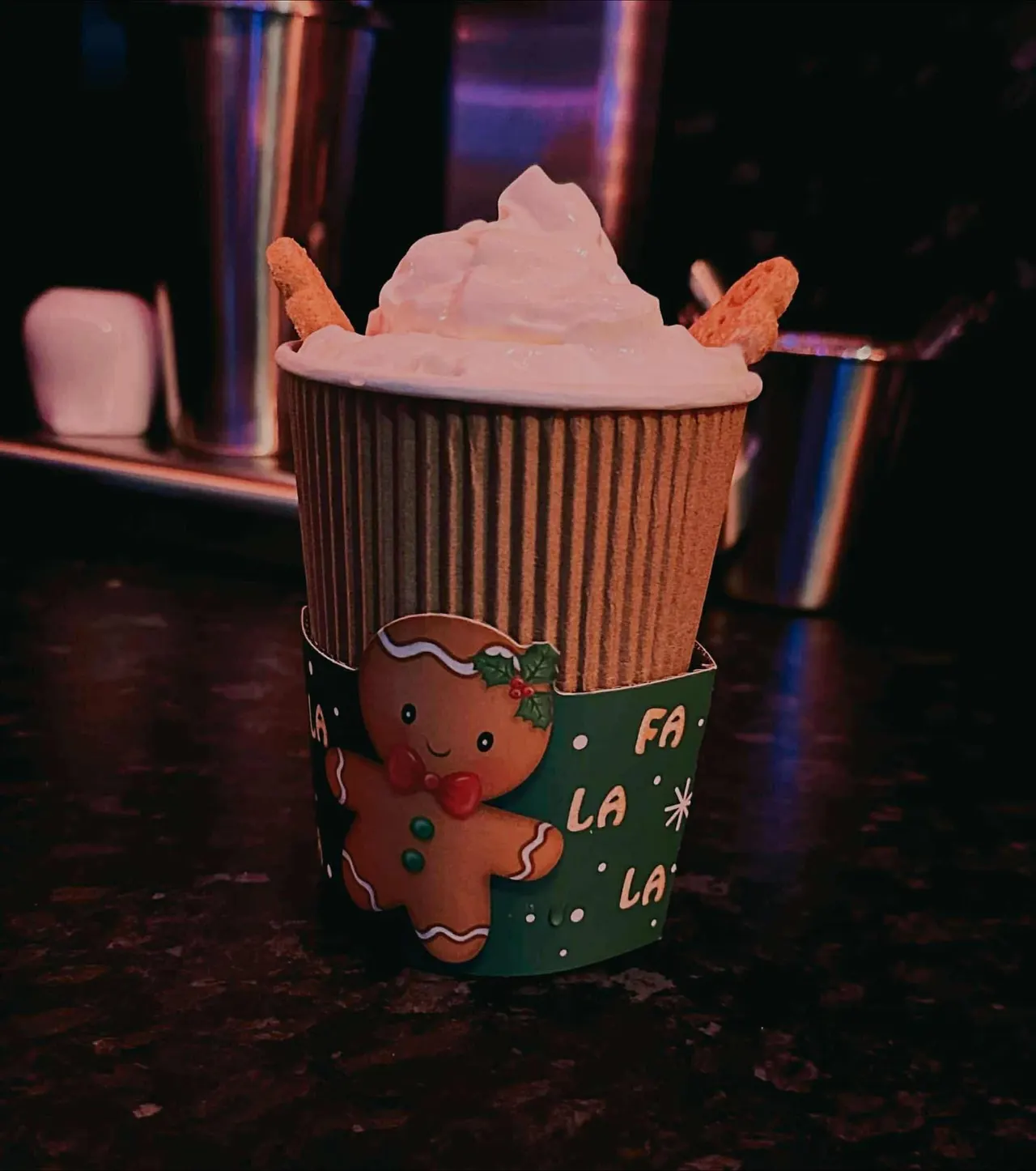 Festive cupcake with pink frosting and biscuit decoration in gingerbread-themed wrapper, lit by coloured neon lighting.