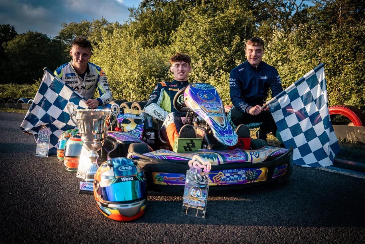 Three young kart racers pose with chequered flags and trophies after a race at an outdoor karting track.