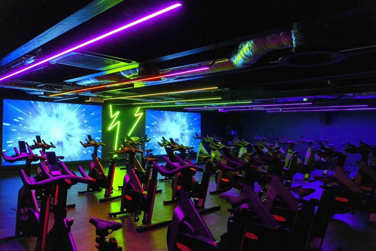 Indoor cycling studio with rows of exercise bikes lit by neon purple, blue and green lights and screens displaying workout visuals.