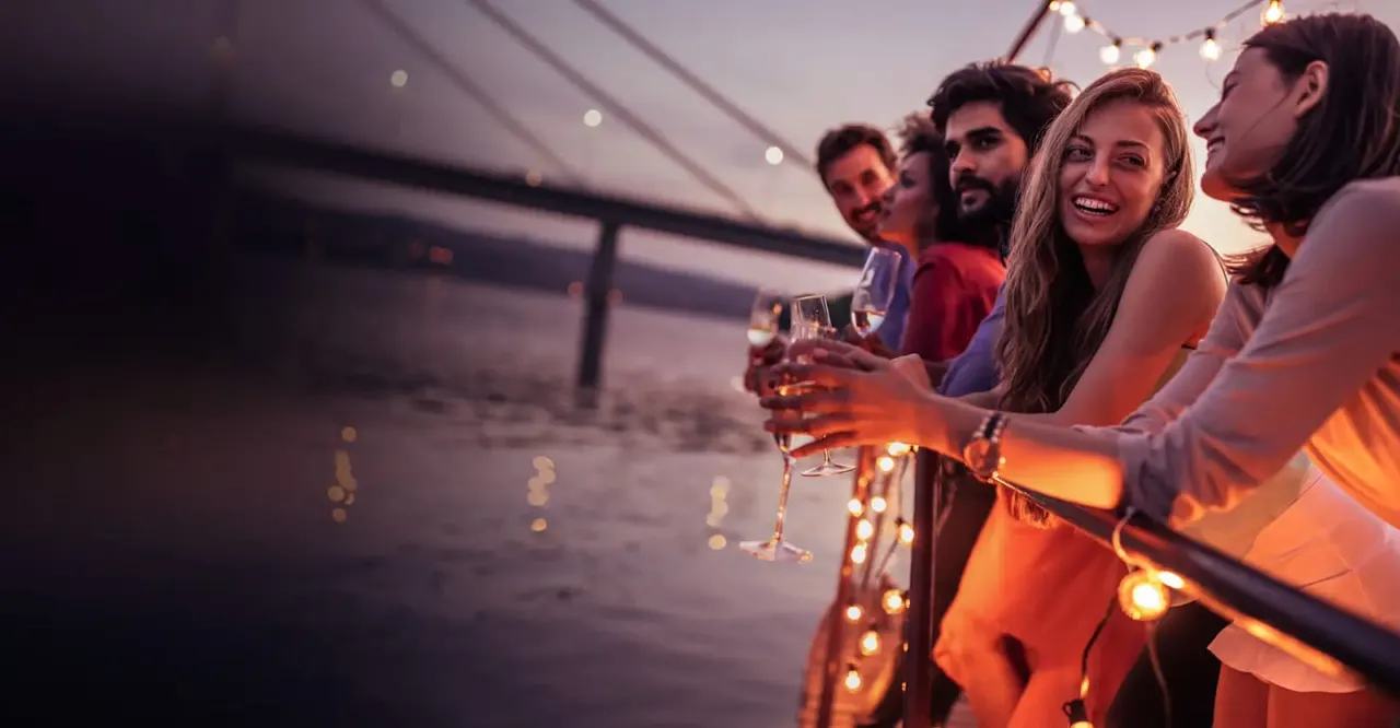 Group of friends holding drinks on a boat deck at sunset with string lights overhead.