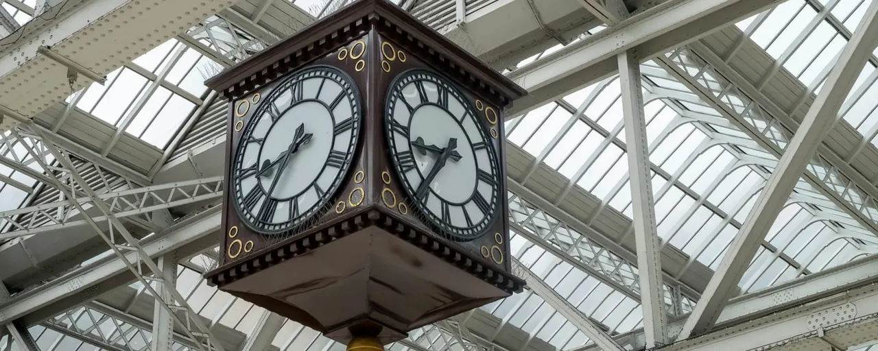 Ornate four-faced clock hanging from a Victorian iron and glass station roof structure.
