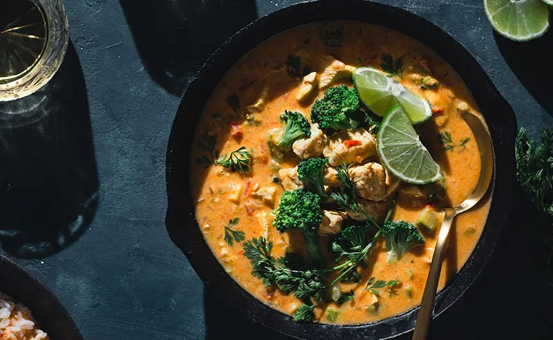 Overhead view of a creamy curry with broccoli and lime in a black bowl on a dark surface.