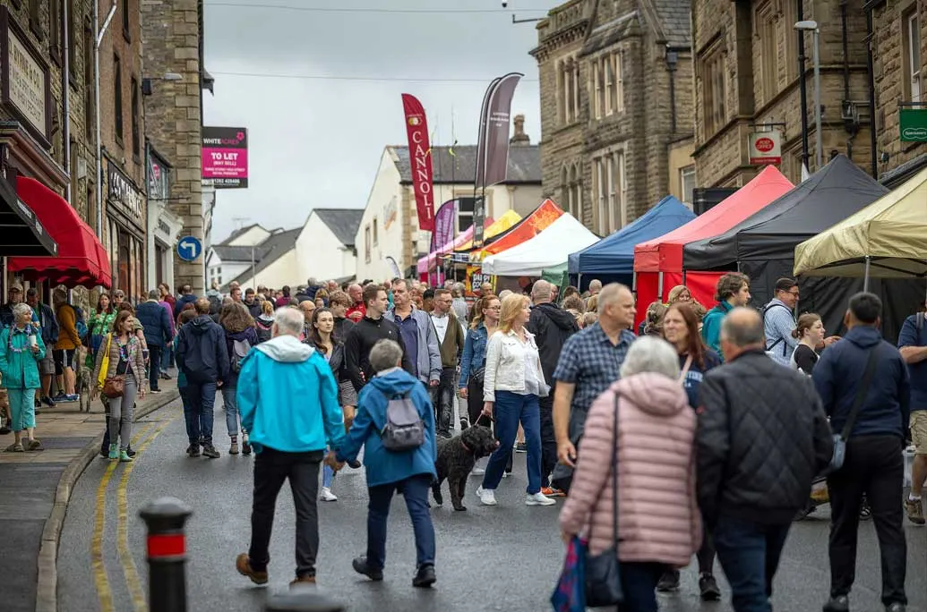 Busy outdoor street market with crowds of visitors walking past colourful market stalls and tents lining a town centre street.