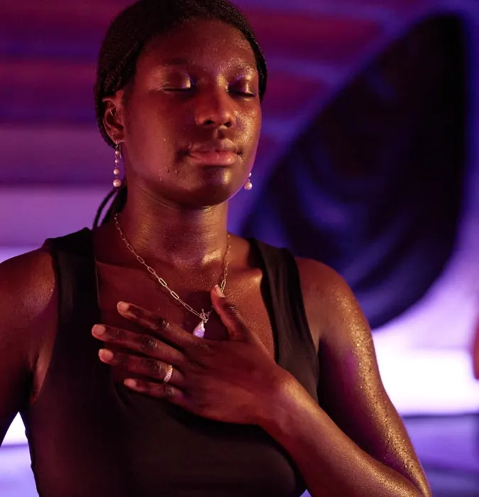 Woman in a black tank top with eyes closed, hand on chest, in purple and red stage lighting.