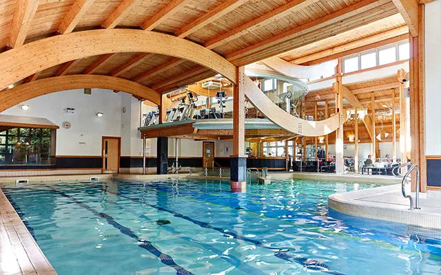 Indoor swimming pool with lane dividers, wooden arched ceiling, spectator gallery, and water slide visible in bright, modern leisure facility.