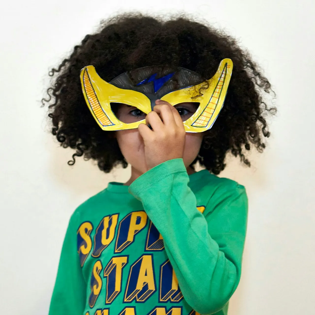 Child wearing a yellow superhero mask and green shirt, holding the mask to face against white background.