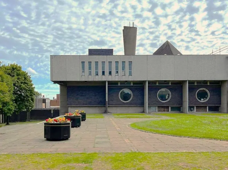 Modern brutalist library building with circular porthole windows and entrance courtyard with planted bollards.