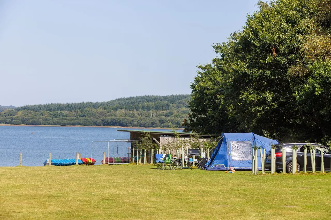 Lakeside campground with tent, vehicles, and water sports equipment on grassy field overlooking forested hills.