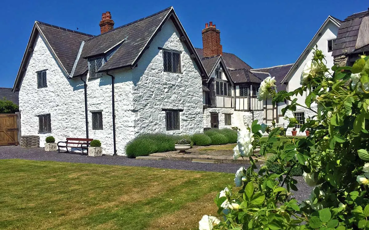 Historic whitewashed stone cottage with slate roof and brick chimneys, surrounded by manicured gardens and lawn under clear blue sky.
