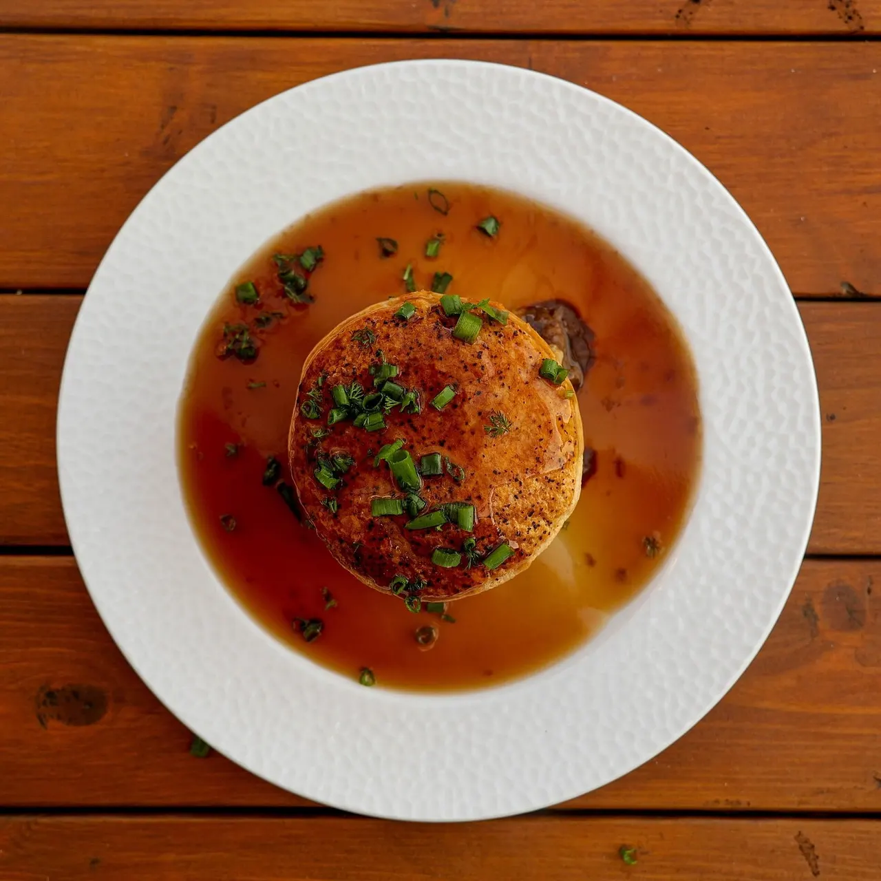 Seared scallop in amber broth garnished with green onion, served on a white plate on wooden table.