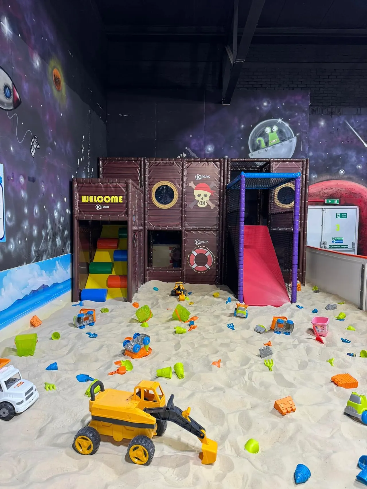 Indoor play area with sand pit containing toy vehicles and colourful play equipment, themed pirate ship structure with slides and climbing features behind.