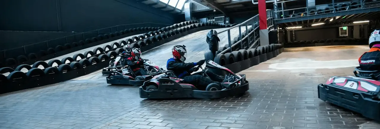 Multiple go-kart racers navigate an indoor track lined with black tyres, wearing helmets and racing suits.