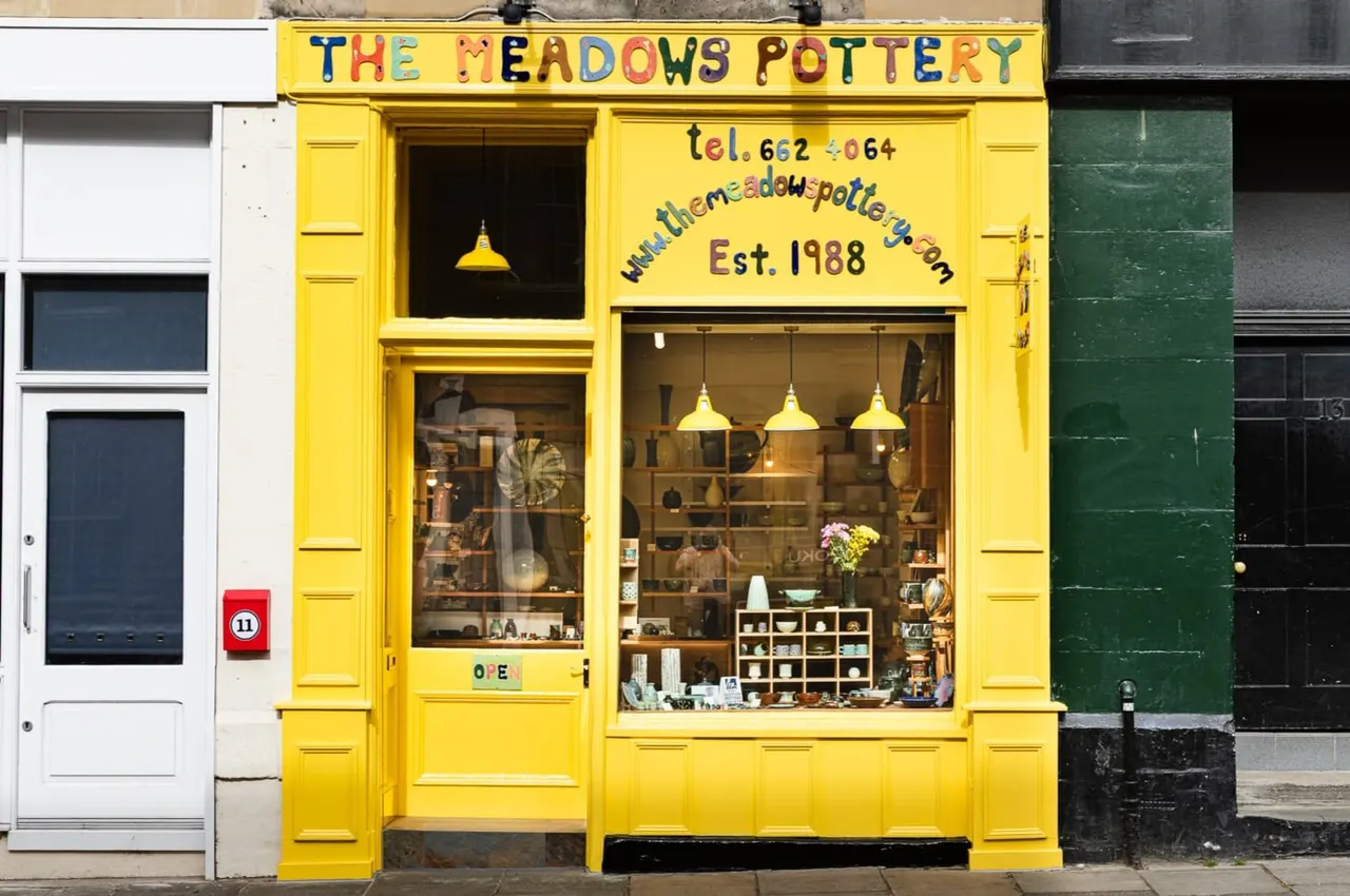 Bright yellow pottery shop storefront with yellow pendant lights, shelves displaying ceramic items, and flowers in the window.
