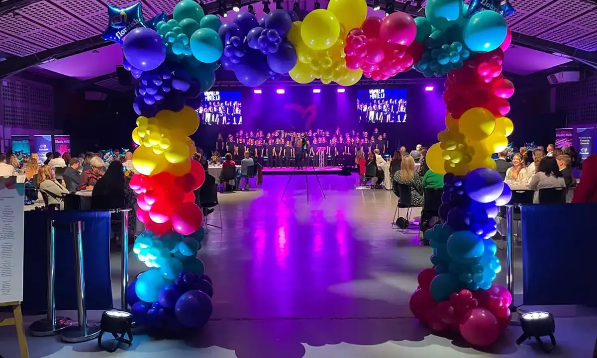 Indoor event space with colourful balloon installations, seated guests, and a stage with performers under purple lighting.