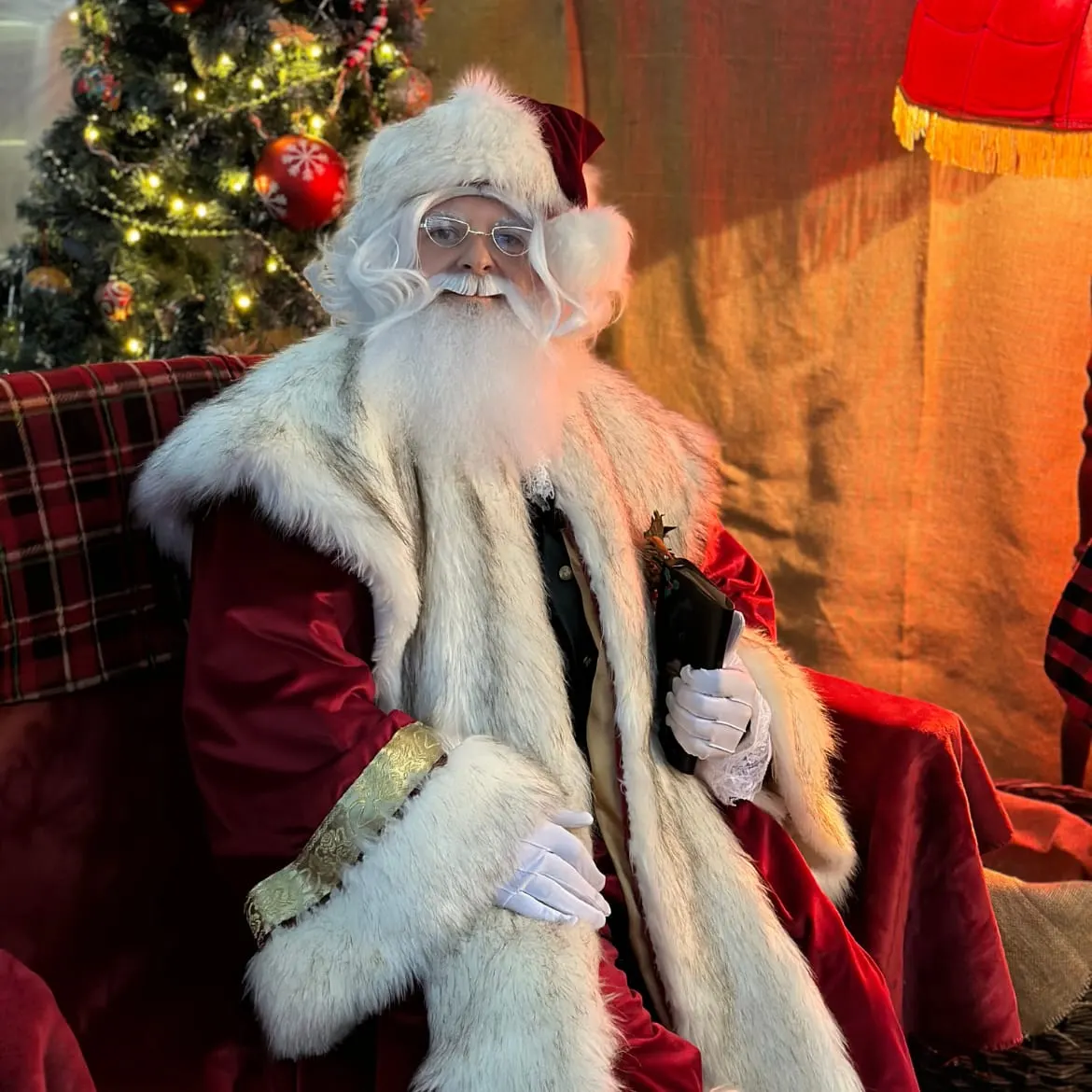 Person dressed as Father Christmas seated in festive setting with decorated Christmas tree and red furnishings.