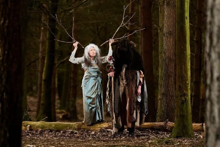 Two performers in theatrical costume stand in a dense woodland, one holding bare branches aloft.