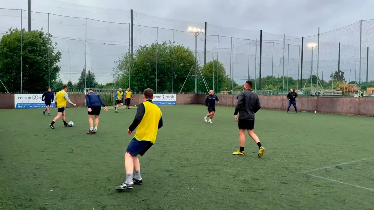 Players in yellow and navy bibs competing in a football match on an artificial pitch at dusk with floodlights.