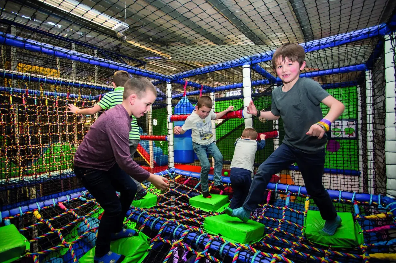 Children playing and jumping in an indoor soft play area with coloured netting, foam obstacles and trampolines.