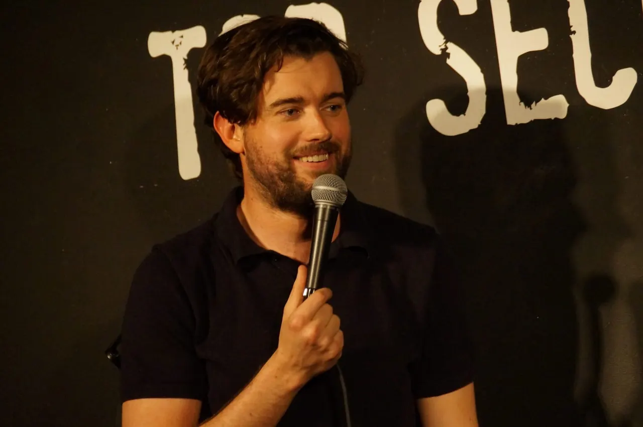 A man in a black shirt holds a microphone whilst performing on stage at a comedy club.
