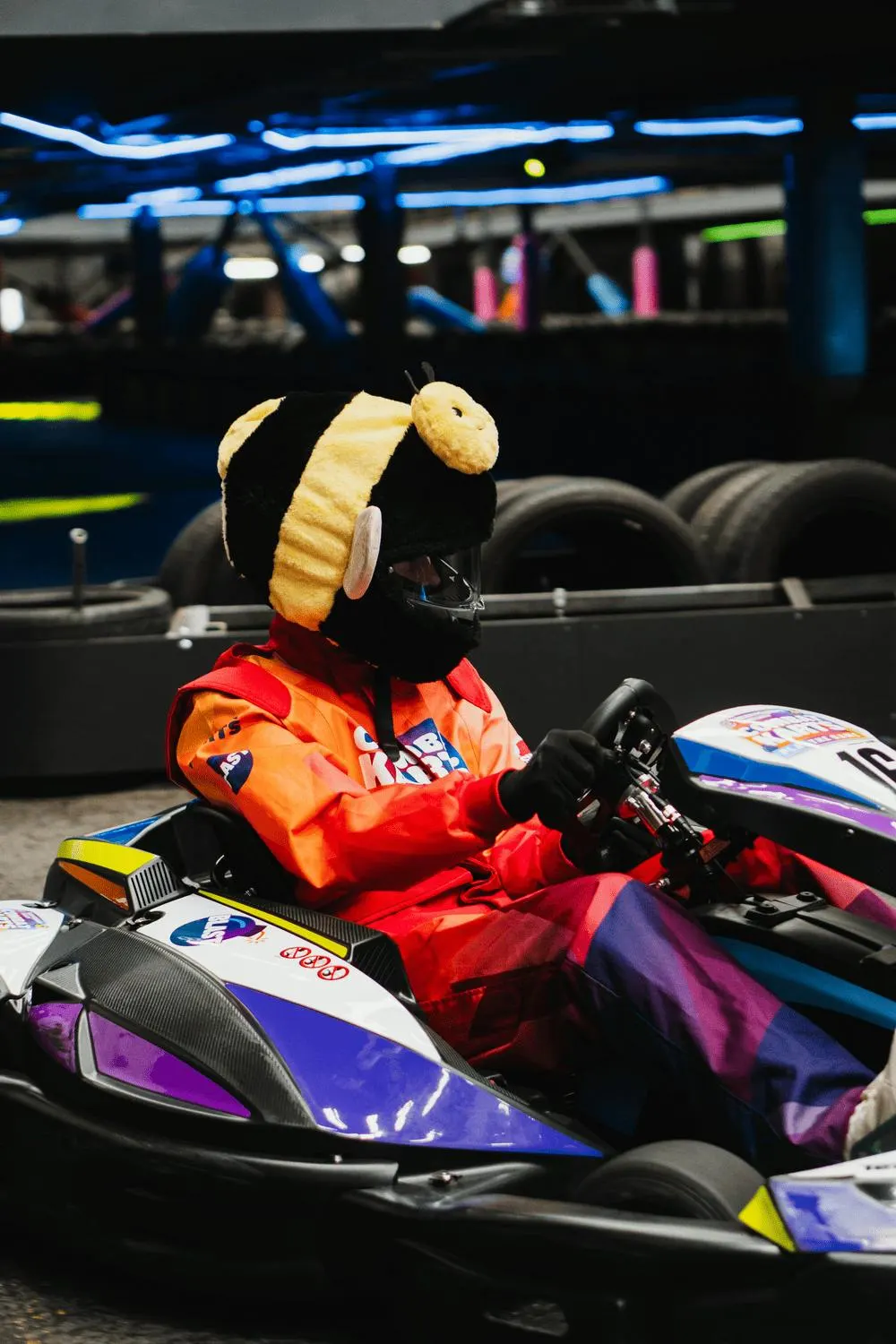 Costumed mascot character seated in a go-kart at an indoor karting venue with neon lighting and tyres visible in background.