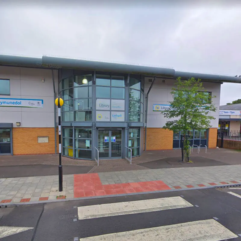 Modern community hub building with glass entrance doors, brick and rendered exterior, and paved forecourt under cloudy sky.