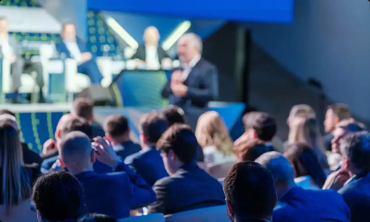 An audience seated in blue chairs watches a presenter on stage at an indoor event.