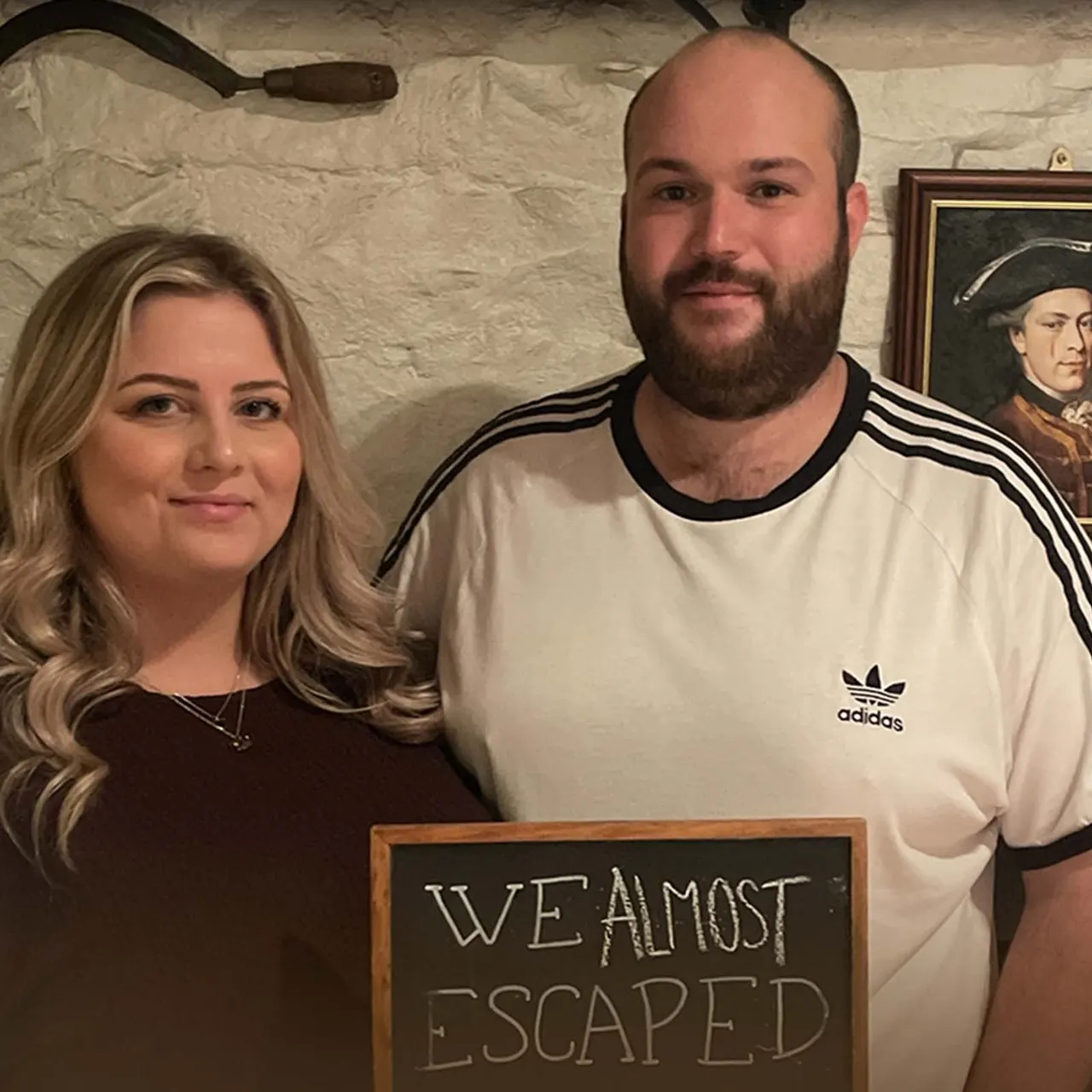 Two people posing indoors against a stone wall, holding a chalkboard sign reading "We Almost Escaped".