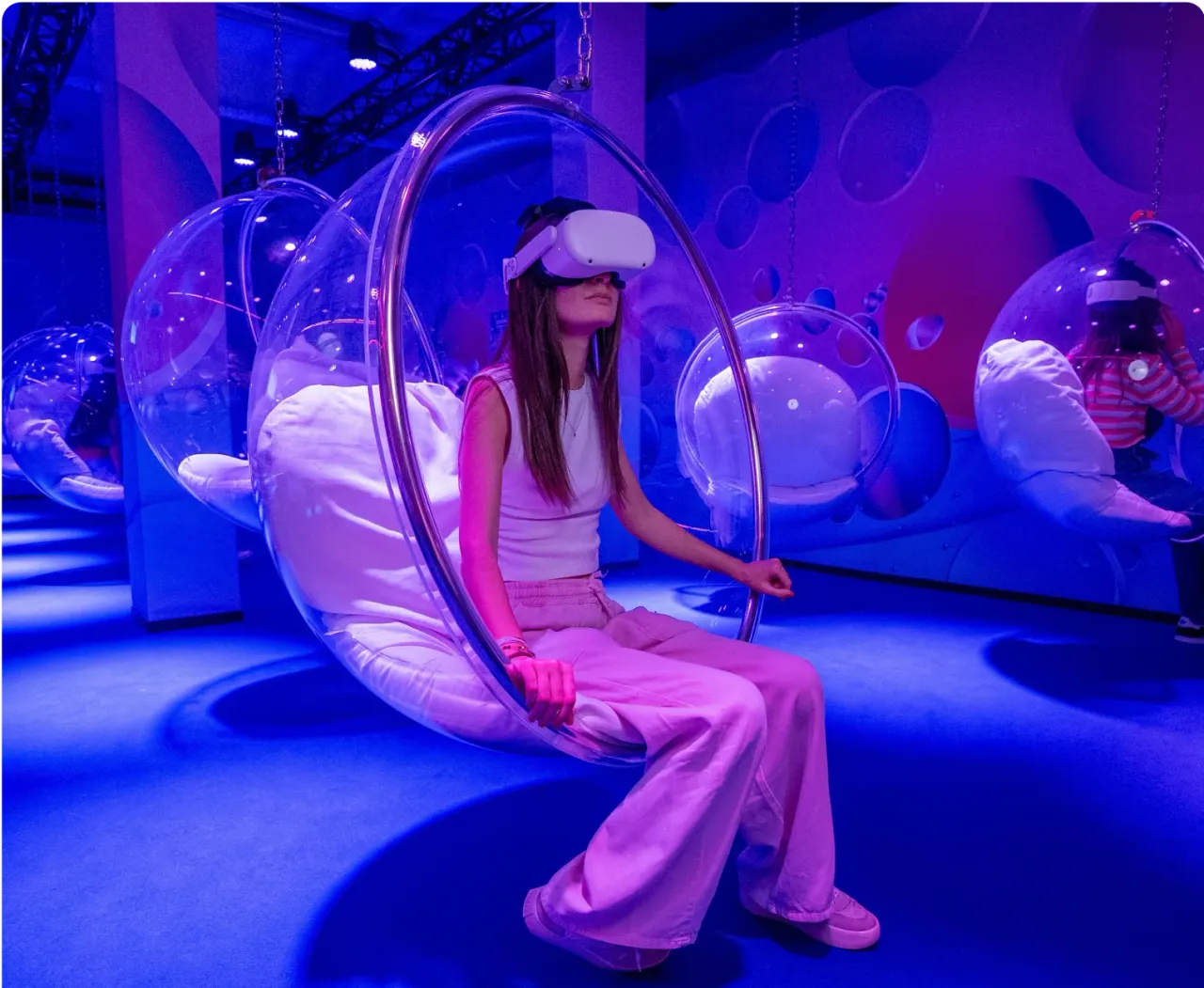 Woman wearing a VR headset sits in a transparent bubble pod suspended from the ceiling in a neon-lit purple and blue room.