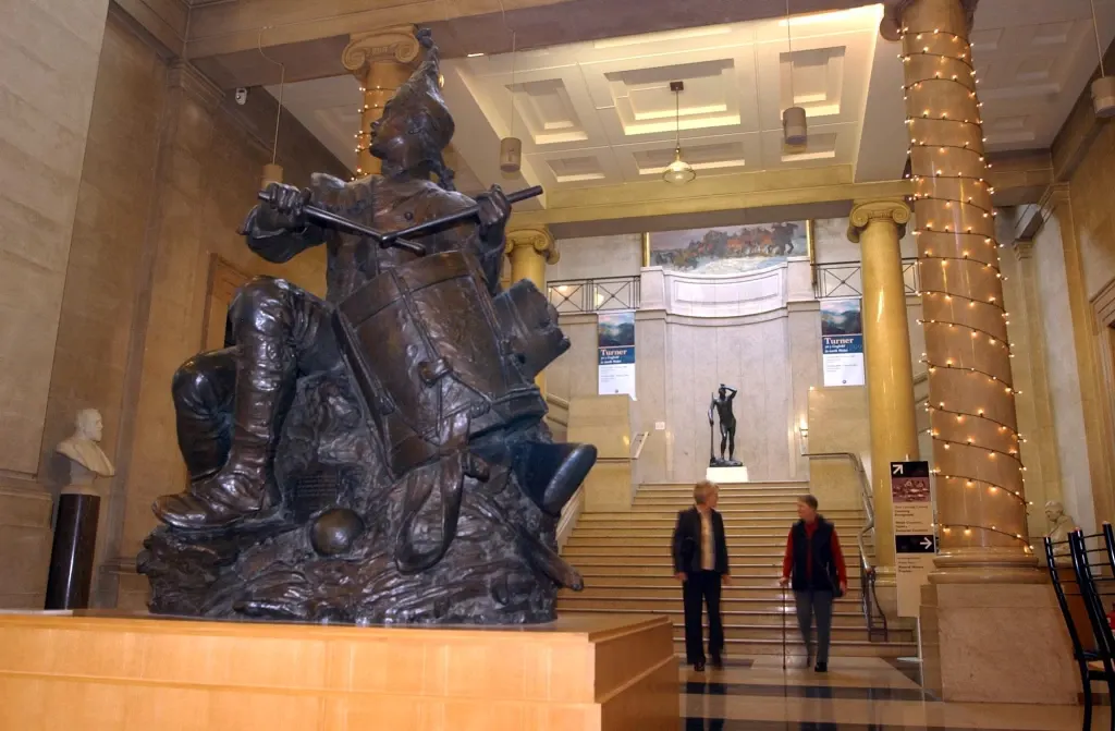 Grand museum hall with large bronze equestrian statue, two visitors standing near ornate staircase with decorative columns.