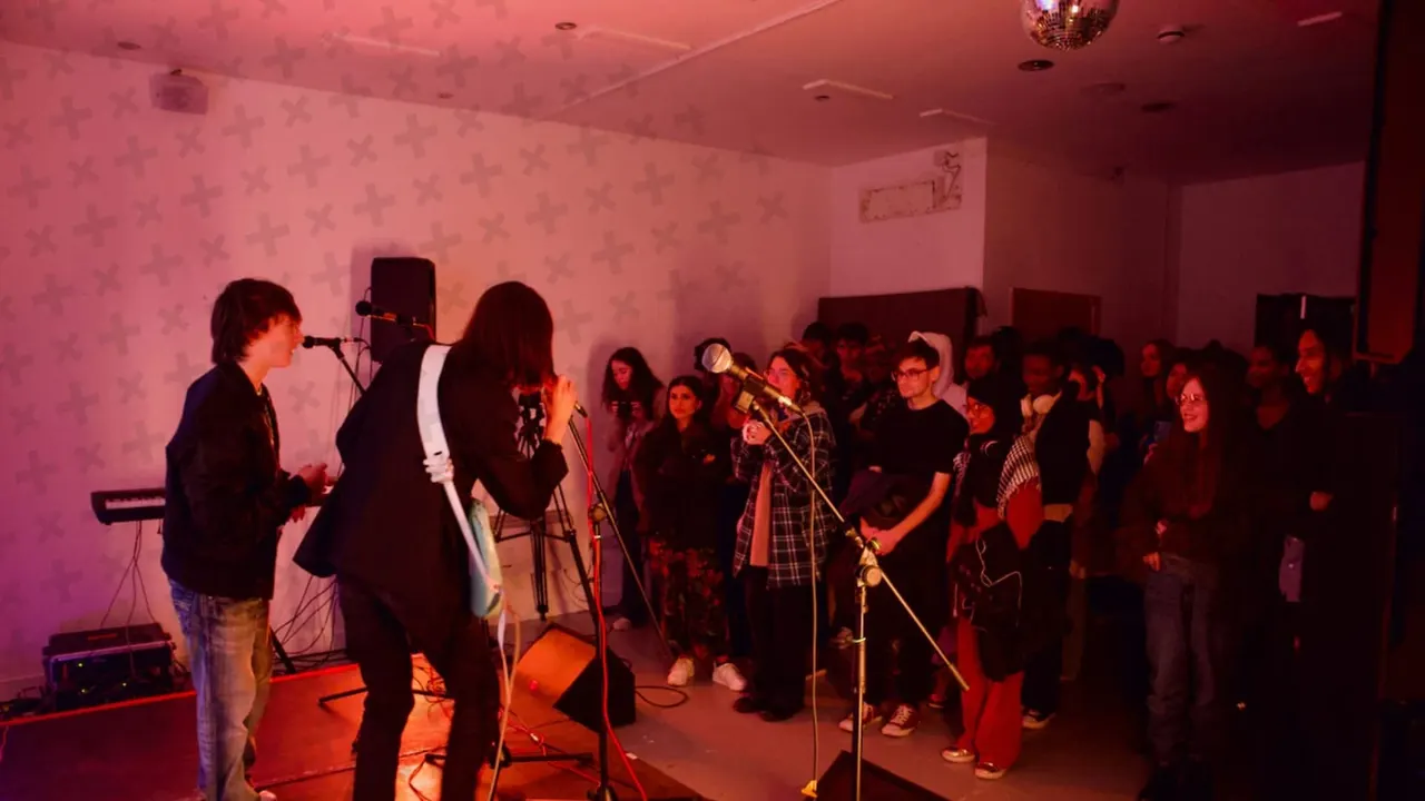 Band performing on stage with red and pink ambient lighting, audience gathered around watching the live music performance.