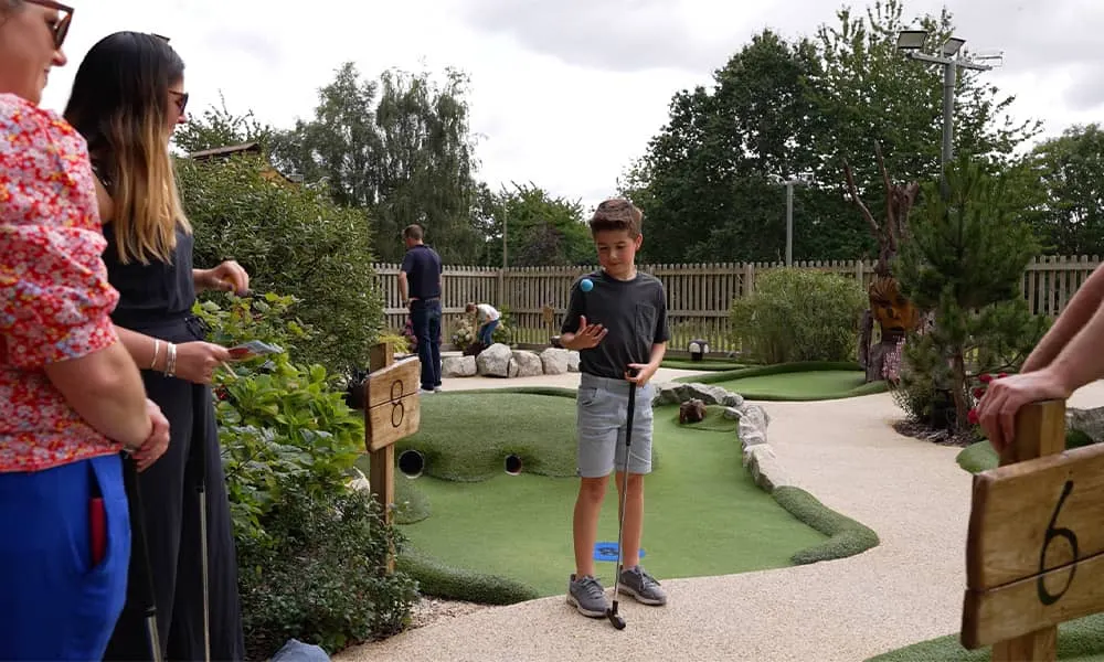 Children and adults playing mini golf on an indoor course with artificial turf and wooden markers.
