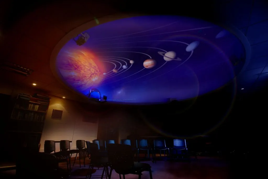 Planetarium dome display showing solar system with sun and planets whilst visitors sit in tiered seating below.