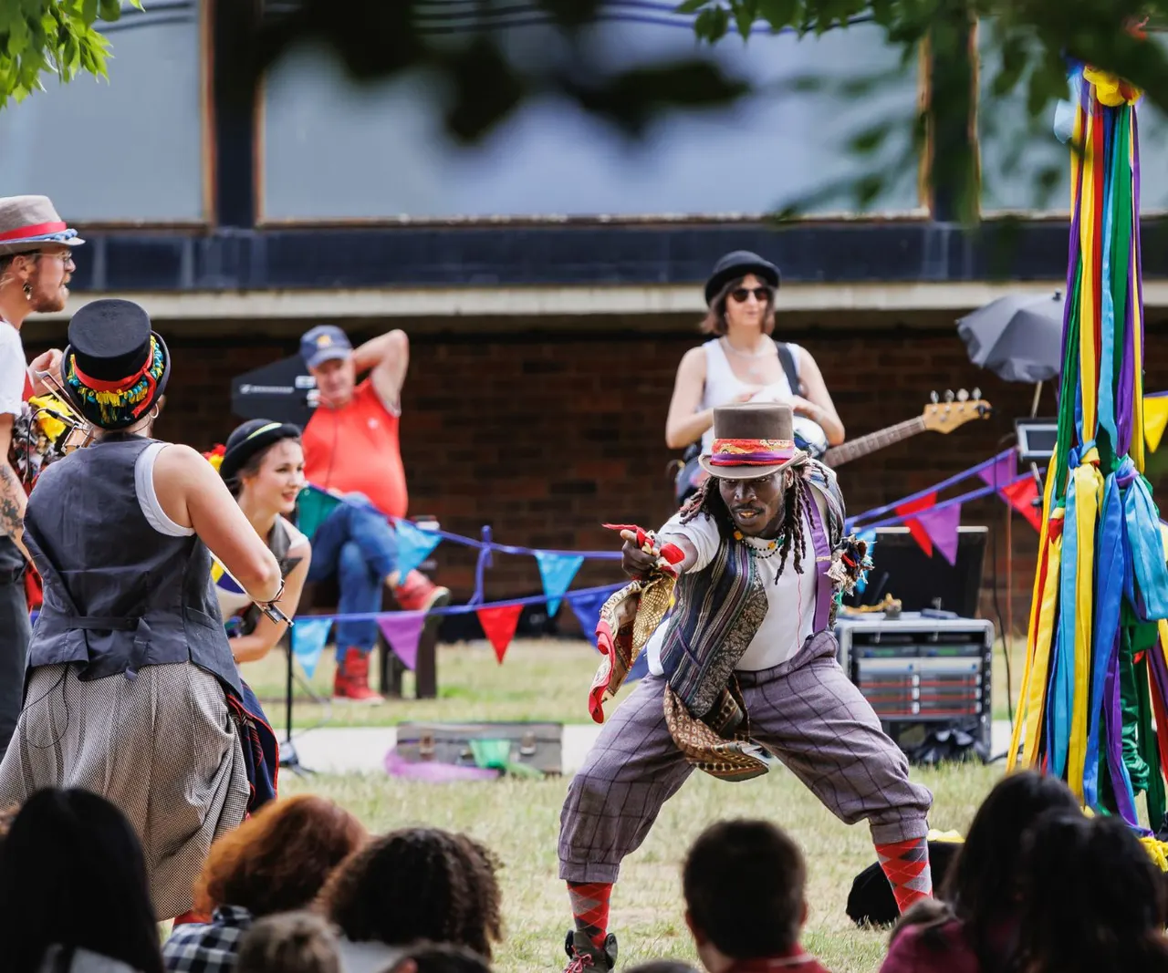 Colourfully dressed performers entertain a crowd at an outdoor festival event with bunting and rainbow decorations.