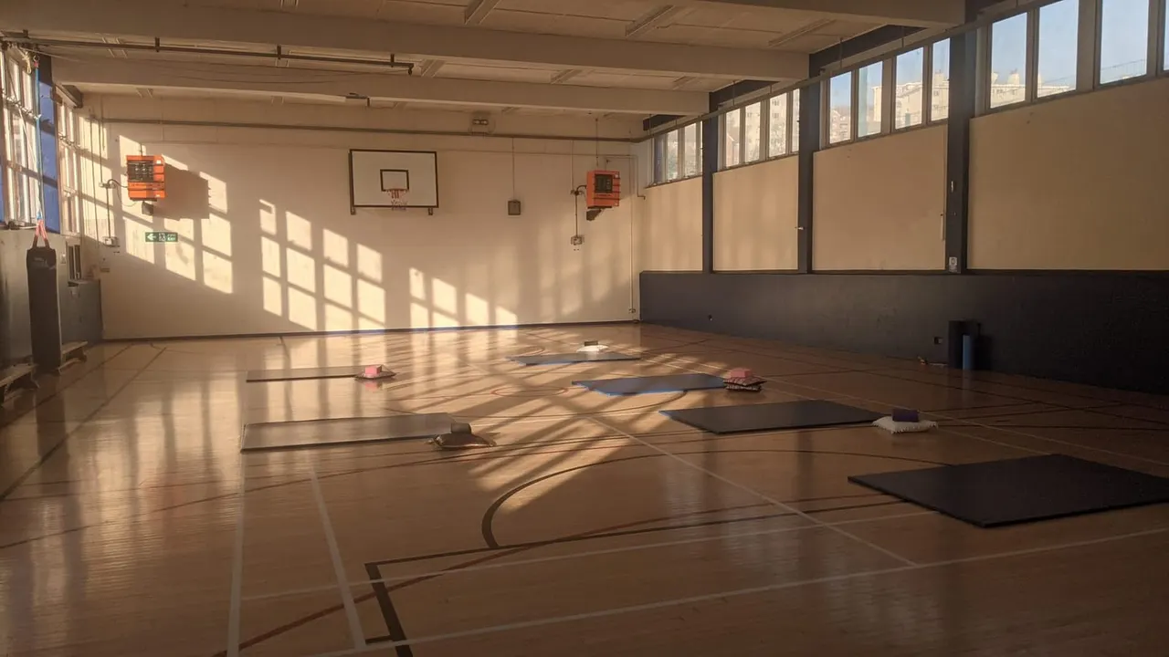 Empty indoor gymnasium with wooden floor, basketball hoops, exercise mats and natural light from large windows.