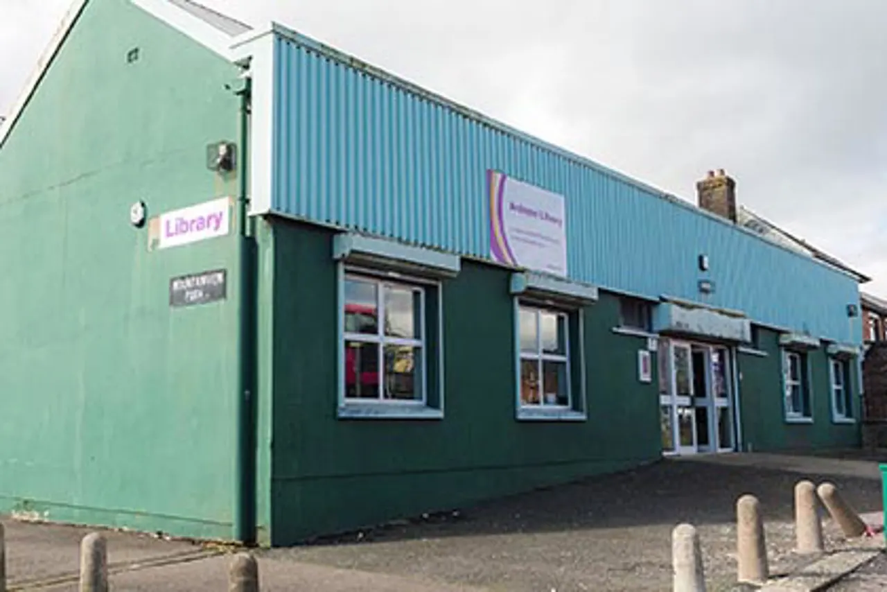 A modern single-storey building with green and blue corrugated metal cladding, featuring multiple windows and entrance doors, with a Library sign visible on the left side.