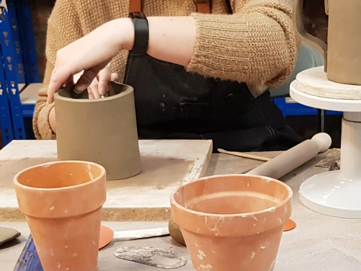 Person in beige jumper working with clay at a pottery wheel, surrounded by terracotta pots and ceramic tools.