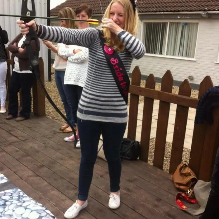 Woman aiming a bow and arrow whilst others watch at an outdoor activity venue.