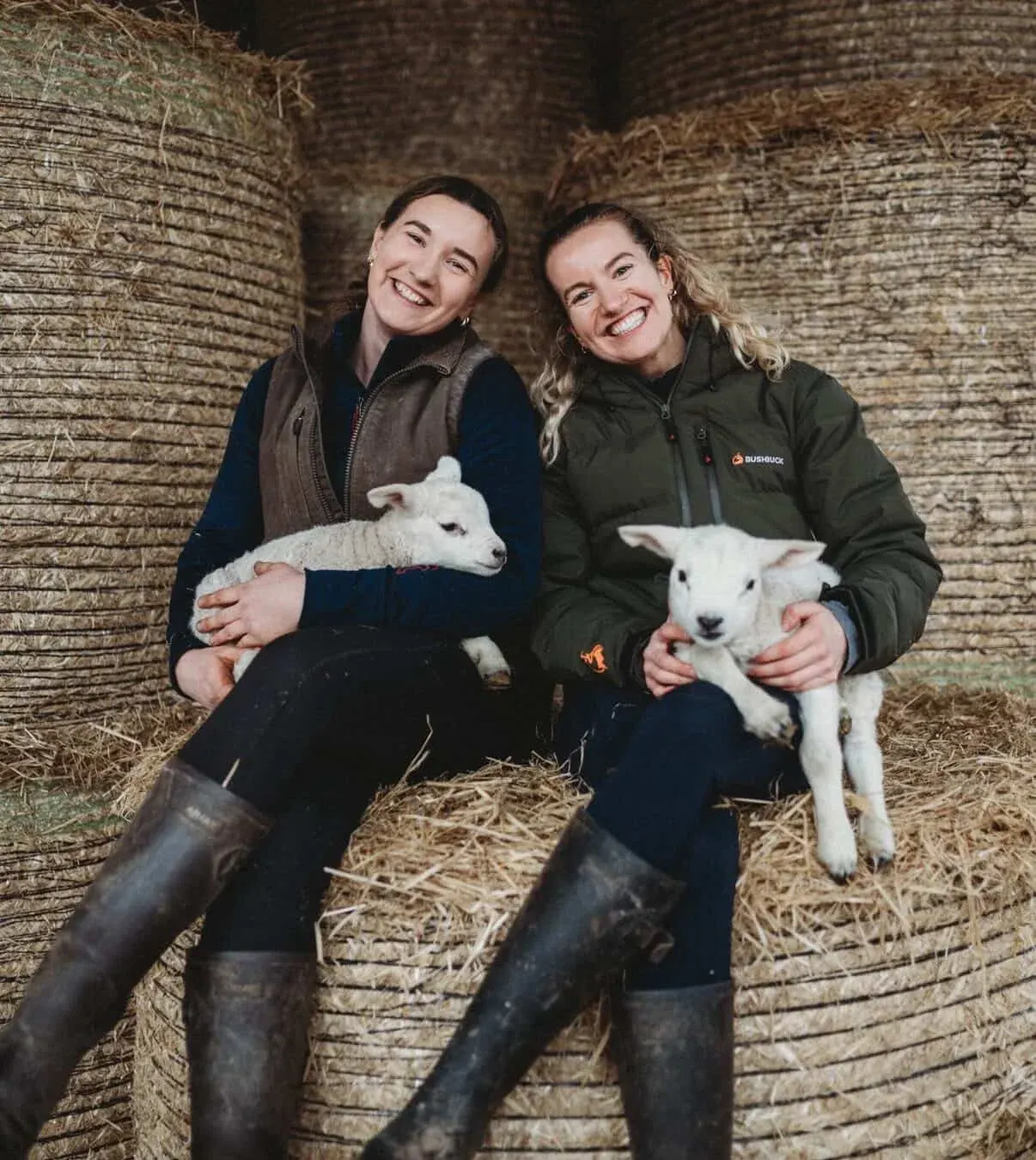 Two smiling women holding young lambs in a barn with hay and wooden fencing.