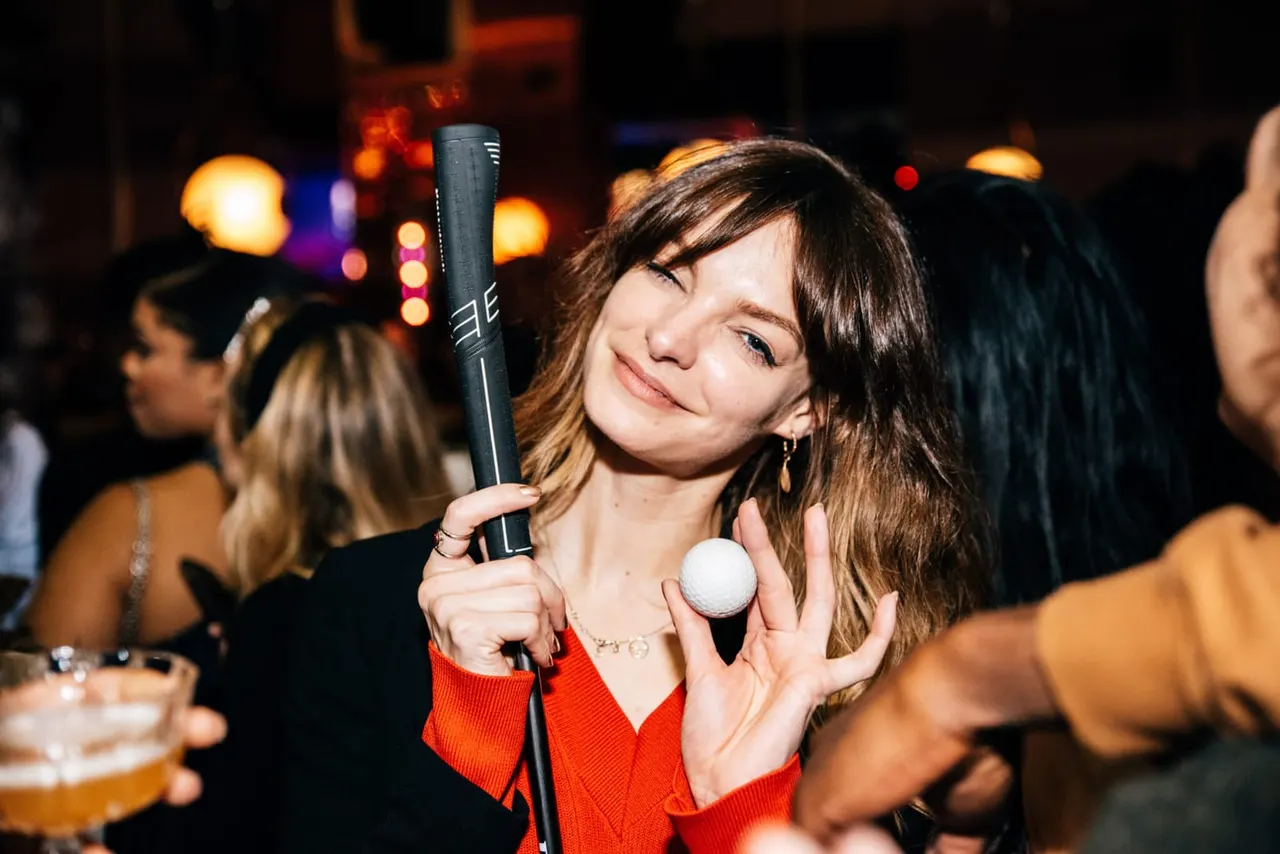 Woman holding a golf club and ball at an indoor golf venue during an evening event with warm ambient lighting.