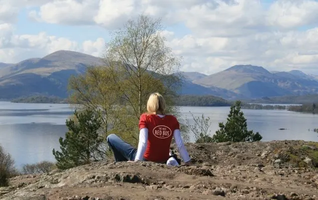 Person sitting on rocky ground overlooking a loch with mountains in the background.