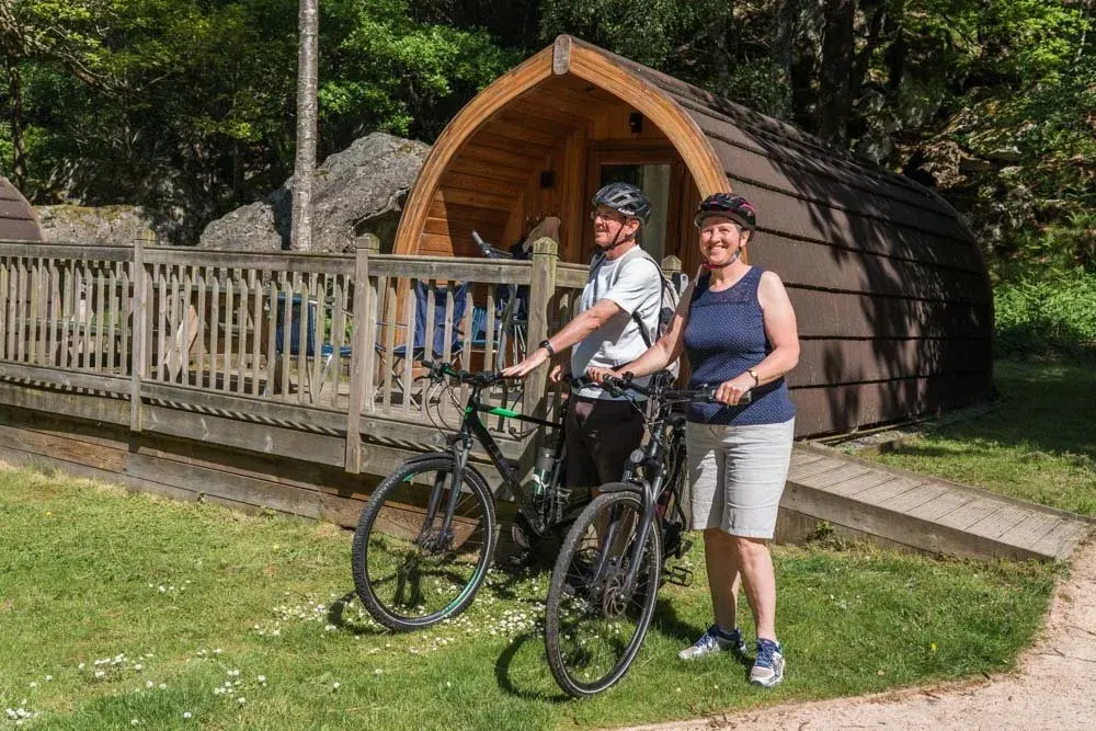 Two cyclists with helmets stand with their bikes on grass outside a wooden barrel-shaped glamping pod surrounded by trees.