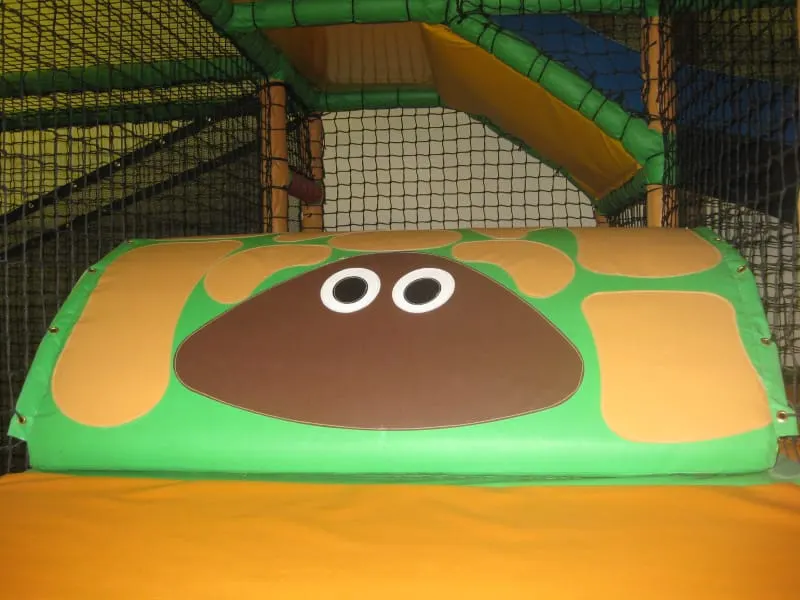 Colourful soft play equipment featuring a green and brown character face inside an indoor play structure with netting and slides.