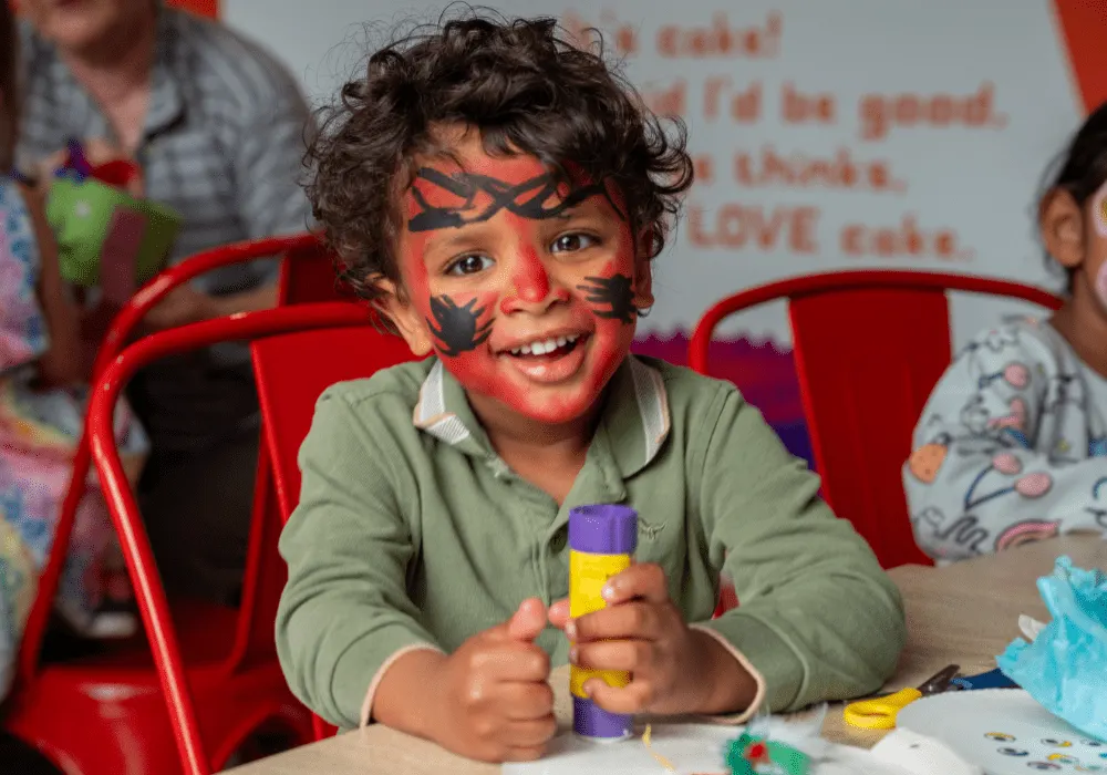 Young child with face paint smiling at table with craft supplies during an activity session.
