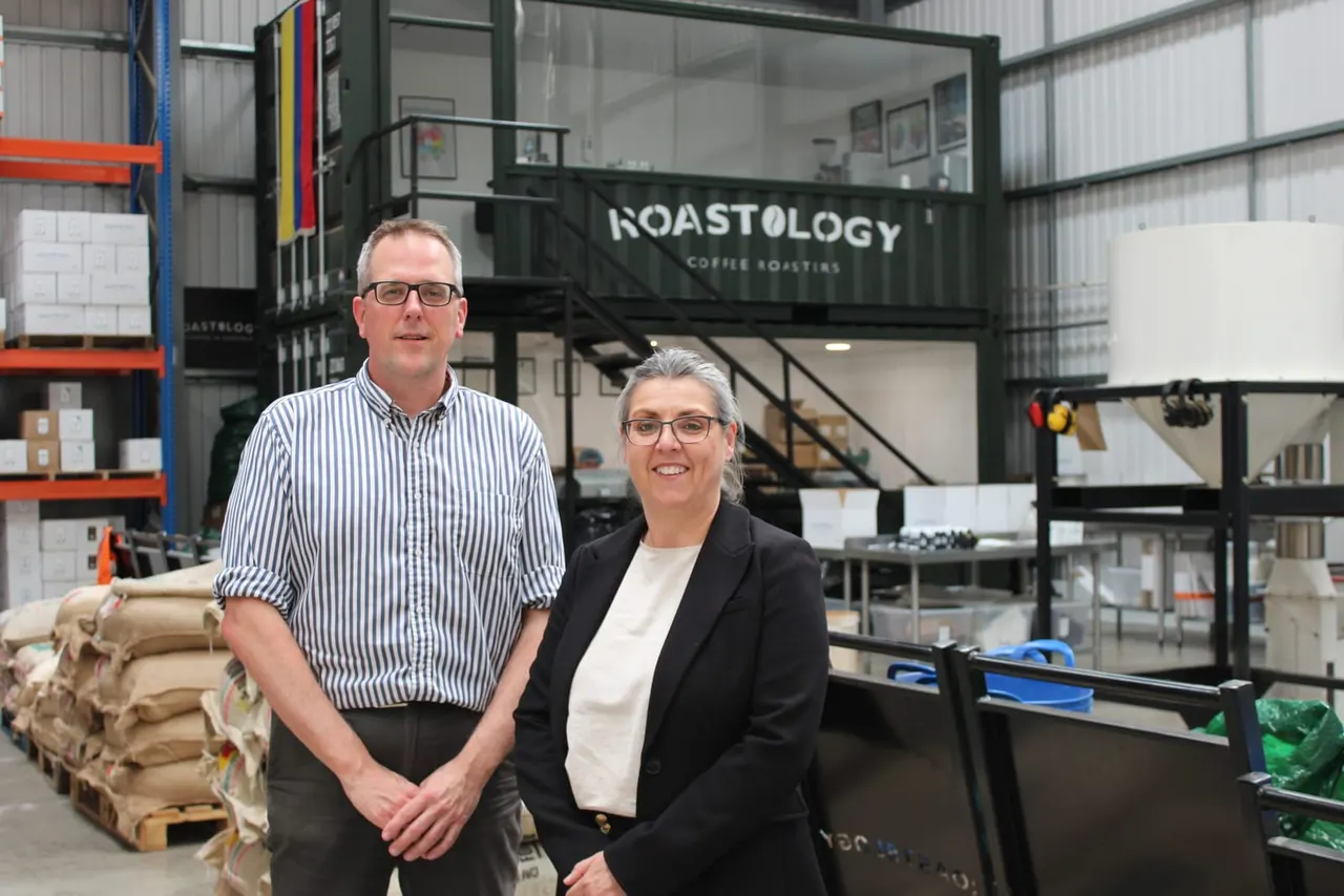 Two people stand in front of a Roastology coffee roastery building inside an industrial warehouse space.