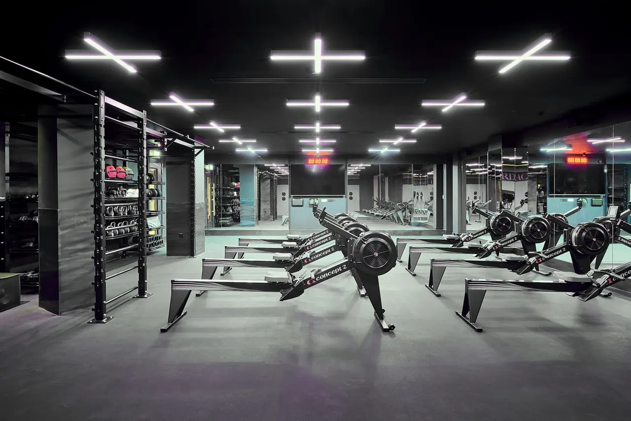Modern gymnasium interior with rowing machines arranged in rows under neon lighting and mirrored walls.